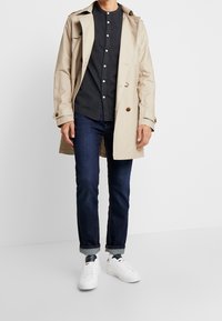 Cappotto trench beige sopra una camicia scura con bottoni, jeans blu e sneakers bianche. Presenta bottoni marroni e orli risvoltati sui jeans.