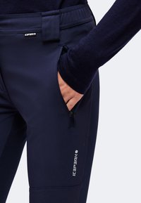 Main dans la poche zippée d'un pantalon softshell bleu marine foncé avec des logos Icepeak à la taille et sur la cuisse.