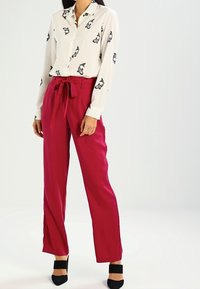 Blouse blanche à imprimé animalier noir, associée à un pantalon taille haute, jambes larges, rose vif, noué à la taille. Des talons noirs complètent la tenue.