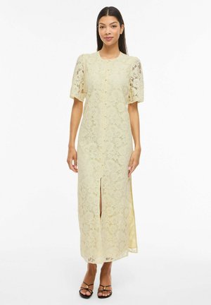 Femme portant une robe midi en dentelle florale jaune pâle, à manches courtes, fente sur le devant, et des talons ouverts noirs, debout sur fond blanc.