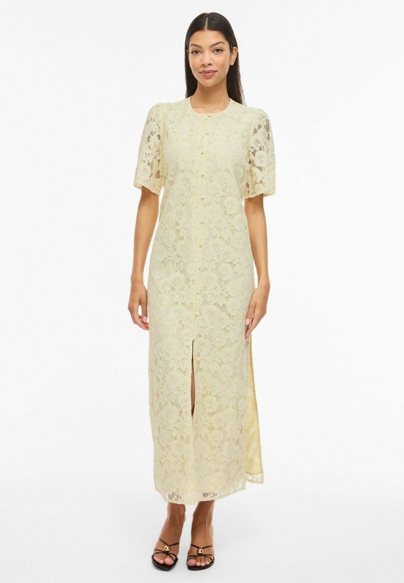 Femme portant une robe midi en dentelle florale jaune pâle, à manches courtes, fente sur le devant, et des talons ouverts noirs, debout sur fond blanc.