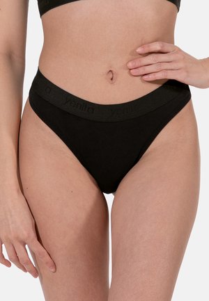 Sezione centrale del corpo di una donna che indossa mutandine nere a vita alta con elastico marchiato, una mano appoggiata sull'anca, su sfondo chiaro.