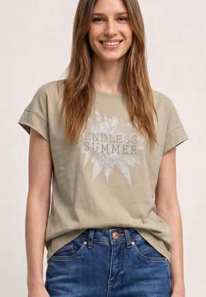 Uśmiechnięta kobieta z długimi brązowymi włosami, ubrana w beżowy T-shirt z napisem „ENDLESS SUMMER” i grafiką kwiatów, zestawiona z niebieskimi dżinsami.