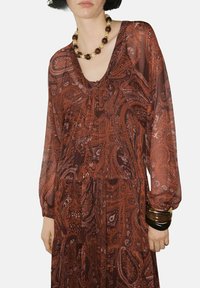 Vestido largo y fluido en un rico tono óxido con patrón de paisley, que presenta mangas largas y escote en V. Los accesorios incluyen un collar grueso y pulseras.