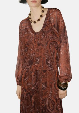 Abito lungo e fluente in un ricco color ruggine con motivo paisley, caratterizzato da maniche lunghe e scollo a V. Gli accessori includono una collana chunky e bracciali.