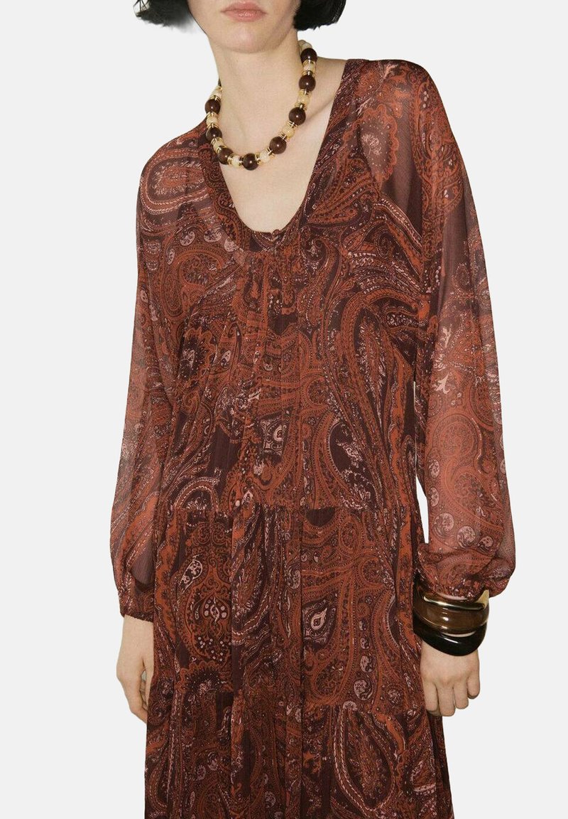 Vestido largo y fluido en un rico tono óxido con patrón de paisley, que presenta mangas largas y escote en V. Los accesorios incluyen un collar grueso y pulseras.