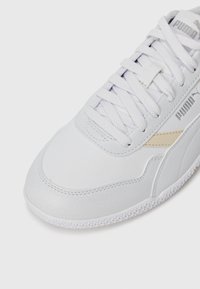 Baskets blanches en cuir synthétique, avec des accents texturés, un système de laçage et un discret logo beige sur le côté.