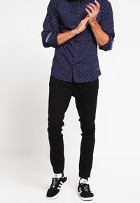 Navyblauwe langemouwen overhemd met witte stippen, gecombineerd met zwarte taps toelopende broek en zwarte sneakers met witte strepen.