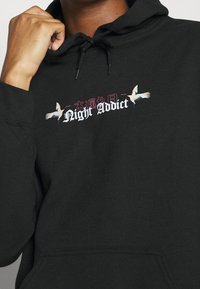 Sudadera negra con bolsillo canguro, que presenta un diseño gráfico de grúas y "Night Addict" en texto blanco en negrita. Capucha con cordón ajustable.