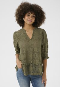 Olīvu zaļas krāsas mežģīņu blūze ar V-veida kakla izgriezumu, trīs ceturtdaļu piedurknēm, ziedu rakstu un volānu aprocēm, apvienota ar ziliem džinsiem.