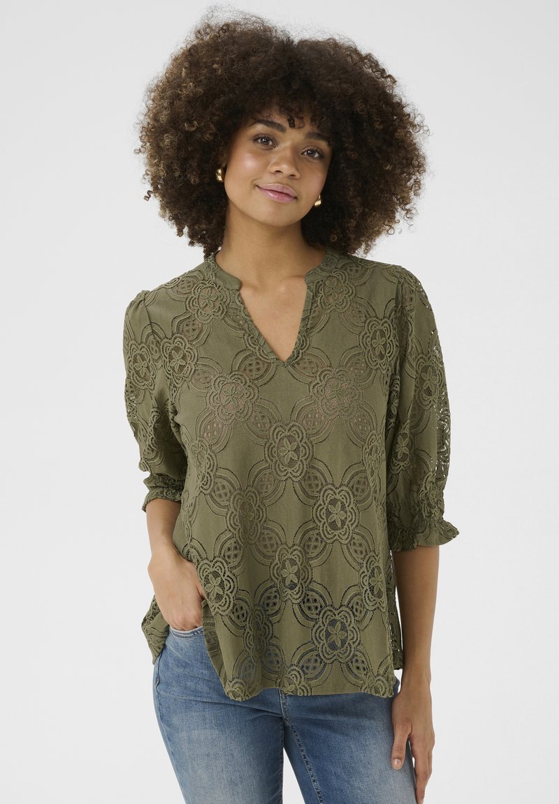 Olīvu zaļas krāsas mežģīņu blūze ar V-veida kakla izgriezumu, trīs ceturtdaļu piedurknēm, ziedu rakstu un volānu aprocēm, apvienota ar ziliem džinsiem.