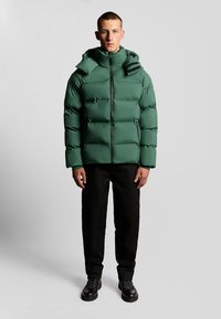 Grön puffjacka med hög krage och justerbar huva, med dragkedjestängning och quiltad textur. Bärs tillsammans med svarta byxor och stövlar.