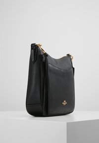 Black leather Coach crossbody bag with gold hardware and tassel detail, displayed on white pedestal against gray background.