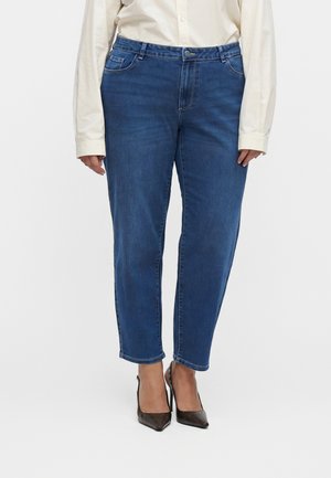 Kvinde iført blå cropped straight-leg jeans, cremefarvet langærmet skjorte og mørke spidse høje hæle, står på hvid baggrund.