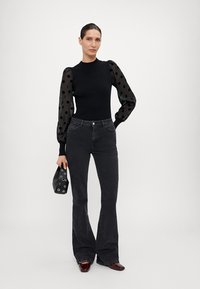 Svart, figurnära topp med genomskinliga prickiga ärmar, matchad med mörka utsvängda jeans. Accessoarerna inkluderar en liten svart väska med silverdetaljer.