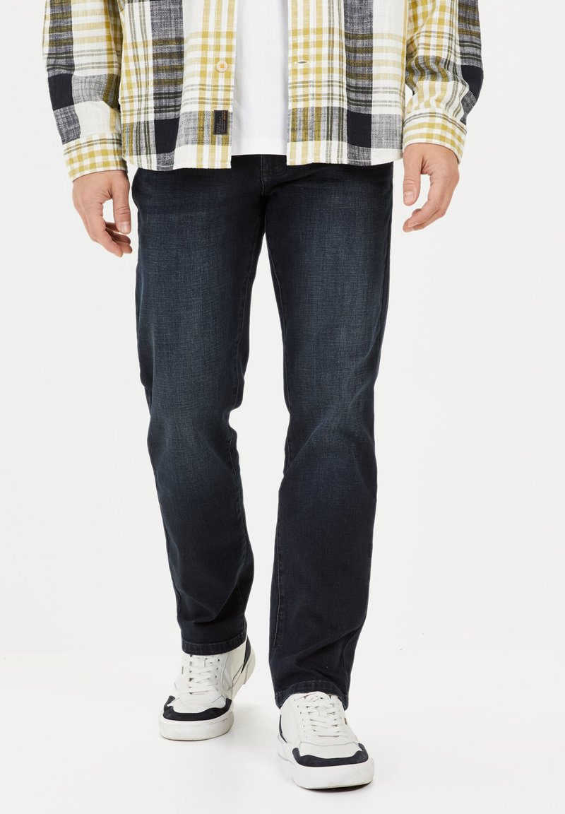 Donkerblauwe spijkerbroek gecombineerd met een geel, zwart en wit geruit overhemd. Het overhemd heeft lange mouwen en knopen, wat textuur aan de outfit toevoegt.