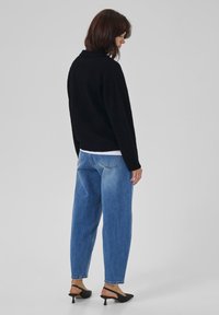 Frau steht seitlich vor einem schlichten Hintergrund, trägt einen schwarzen Pullover, blaue hoch taillierte Jeans und schwarze Slingback-Kitten-Heels.