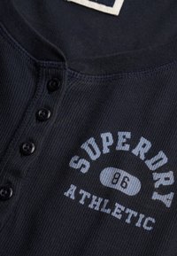 Chemise bleu marine côtelée avec un col henley boutonné et des boutons noirs. "Superdry Athletic" imprimé en blanc et bleu clair sur la poitrine.