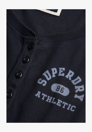 Marineblå ribstrikket skjorte med en henley-krave med knapper og sorte knapper. "Superdry Athletic" trykt i hvid og lyseblå på brystet.
