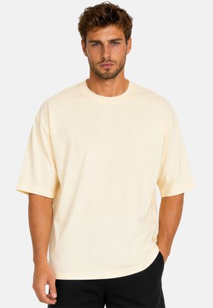 Man met kort bruin haar en baard draagt een losse lichtbeige T-shirt en zwarte broek, staand tegen een effen lichte achtergrond.