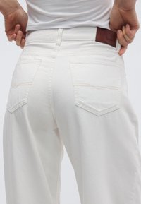 Λευκό τζιν παντελόνι με ψηλή μέση, με πίσω τσέπες που έχουν καμπυλωτές ραφές και καφέ δερμάτινη ετικέτα στη μέση.