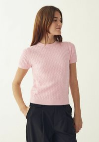 Hele roosa lühikeste varrukatega kootud kampsun, millel on kaabelmustrite mustrid, soonikkoes kaelus ja äär, kombineeritud kõrge vöökohaga mustade pükstega.