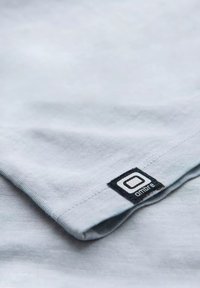Světle šedé tričko s jemnou texturou, na lemu s černou látkovou nášivkou se jménem značky "ombre".