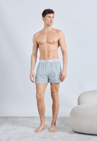 Άνδρας όρθιος, ξυπόλητος μέσα στο σπίτι, φοράει φωτεινά γαλάζια μπόξερ Calvin Klein με μοτίβα, κοιτάζοντας προς τα δεξιά μπροστά σε απλό φόντο.