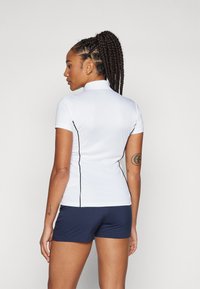 Camicia bianca a maniche corte con colletto alto, con cuciture laterali nere, abbinata a pantaloni corti blu scuro. Materiale liscio ed elastico.