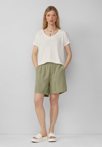 Biała, krótka koszulka z dekoltem w serek, połączona z oliwkowymi szortami z elastycznym pasem oraz białymi sandałami na płaskiej podeszwie. Prosty, casualowy look.