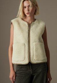 Cremefarvet fleecevest med lynlås foran, rund krave og to forlommer. Kanten er kantet i beige. Ermløs design.