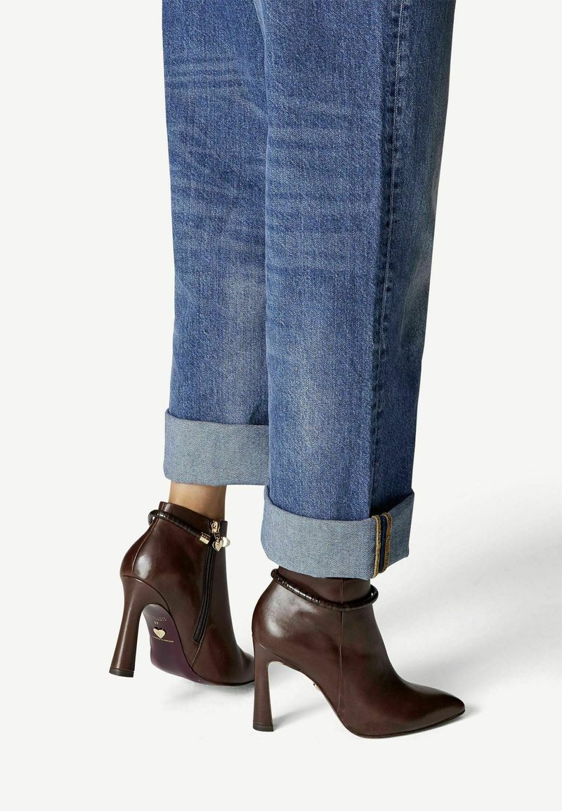 Jean bleu longueur cheville avec larges revers, associé à des bottines en cuir marron foncé à talons hauts, dotées de fermetures éclair latérales et de breloques décoratives.