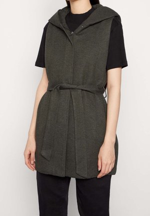 Bezrękawnik w ciemnym oliwkowym kolorze z kapturem, zapinany z przodu na zamek błyskawiczny i z wiązaniem w talii, noszony na czarnej koszulce z krótkim rękawem i czarnych spodniach.