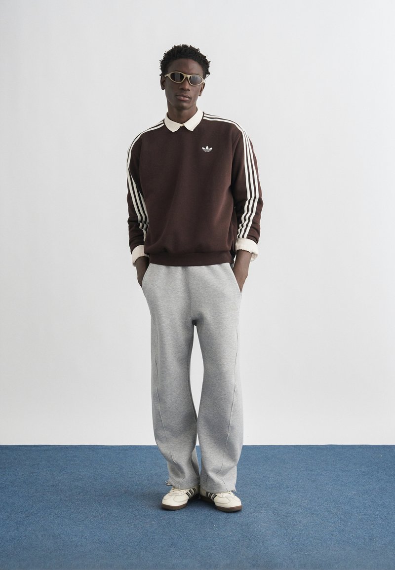 Uomo in piedi con le mani nelle tasche che indossa una felpa Adidas marrone, pantaloni della tuta grigi, scarpe da ginnastica bianche e occhiali da sole su un tappeto blu con sfondo di muro bianco.