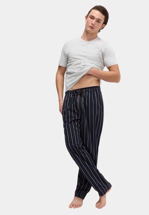 Ung mand løfter hvid T-shirt, iført marineblå pyjamassbukser med striber, stående bosiddende barfodet mod en ensfarvet lys baggrund.