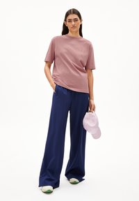 Rosa Baumwoll-T-Shirt mit kurzen Ärmeln, marineblaue weite Hose und eine hellrosa Mütze in der rechten Hand. Weiße Sneakers mit grünen Akzenten.