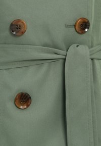 Πράσινο ύφασμα με υφή και διπλό στήθος, με δύο μεγάλα, κυκλικά καφέ κουμπιά και λεπτομέρεια ζώνης από ύφασμα.