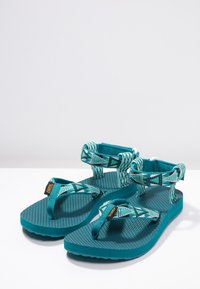 Un paio di sandali da trekking color teal e con motivo, dotati di cinturini regolabili e suole testurizzate, posizionati su una superficie bianca e uno sfondo grigio chiaro.