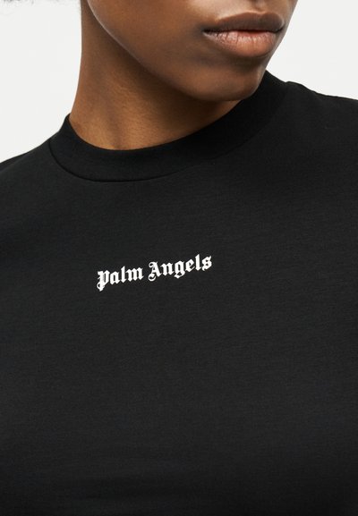 Κοντινό πλάνο προσώπου που φοράει μαύρο μπλουζάκι με στρογγυλή λαιμόκοψη και λευκό λογότυπο "Palm Angels" στο κέντρο του στήθους.
