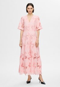 Rochie roz din dantelă cu decolteu în V, mâneci scurte și largi, și închidere cu nasturi. Prezintă modele florale și un tiv în straturi. Purtați-o cu tocuri negre.
