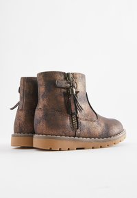 Bottes cheville texturées en bronze avec une fermeture éclair latérale, un détail pompon, et une semelle robuste de couleur beige. Elles présentent un bout arrondi et un design bas.