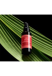 L'huile capillaire multi-usages Aveda Nutri Plenish dans un flacon noir avec une étiquette rose vif, posée sur une feuille de palmier verte sur un fond noir.