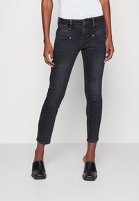 Vaqueros de mezclilla negros con un ajuste slim, diseño de cinco bolsillos, bolsillo delantero con cremallera y costuras de contraste. Combinados con zapatos negros tipo slip-on.
