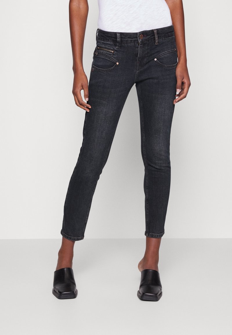 Vaqueros de mezclilla negros con un ajuste slim, diseño de cinco bolsillos, bolsillo delantero con cremallera y costuras de contraste. Combinados con zapatos negros tipo slip-on.
