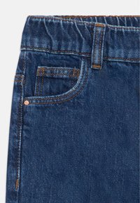 Vaqueros de mezclilla azul oscuro con una cintura relajada, bolsillo frontal con detalle de remaches de cobre y costuras de contraste visibles a lo largo de las costuras.