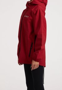 Personne portant une veste à capuche rouge avec le logo "JACKIT", leggings noirs, debout de profil, bras détendus le long du corps sur un fond uni.