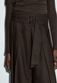 Pantaloni marroni a vita alta con una cintura larga e una fibbia rettangolare, pieghe nette e una texture morbida, che mostrano un taglio su misura.