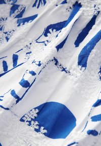 Biała tkanina z niebieskimi kwiatowymi wzorami oraz teksturalnymi białymi detalami haftowanymi. Wzór zawiera duże niebieskie kształty oraz skomplikowane przeszycia.