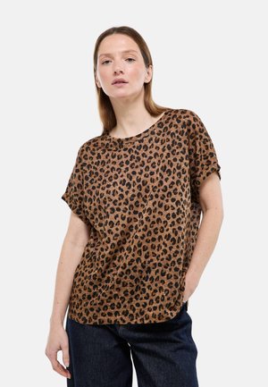 Femme aux cheveux raides châtain clair portant un haut ample à manches courtes imprimé léopard marron et un pantalon foncé, debout avec une main dans la poche.