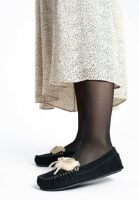 Schwarze Wildleder-Mokassins mit Fellakzenten und beigen Schleifen. Getragen mit durchsichtigen schwarzen Strumpfhosen und einem gemusterten Rock mit asymmetrischem Saum.